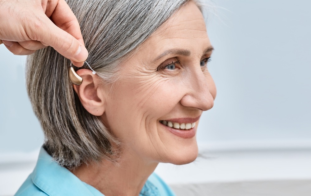 A smiling older woman with gray hair is having a hearing aid placed behind her ear by another persons hand. She appears happy and comfortable.